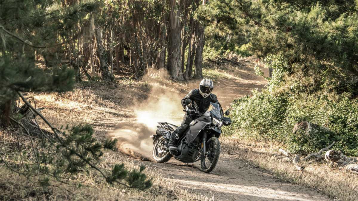 2027 Kawasaki KLR650 DF ABS in Metallic Flat Raw Graystone kicking up dust on a wooded dirt trail