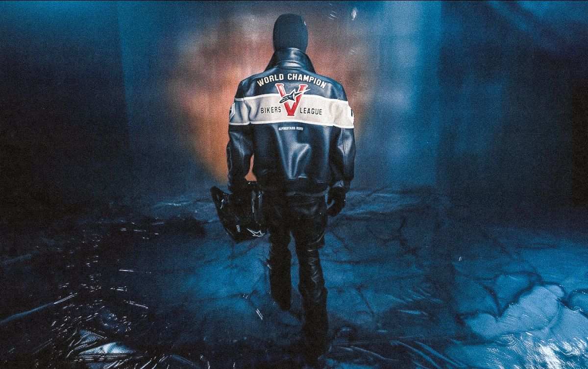 Model walking away from the camera in the Alpinestars RSRV Broken In Varsity Leather Jacket, its back reading World Champion and Bikers League around a red V and Astar logo, carrying a helmet across a moody blue-lit wet floor