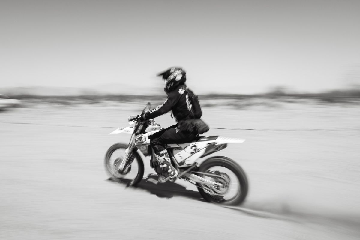 Black-and-white photo of a dirt bike racer speeding across a Johnson Valley dry lake bed, promoting In Dust We Trust 2026.