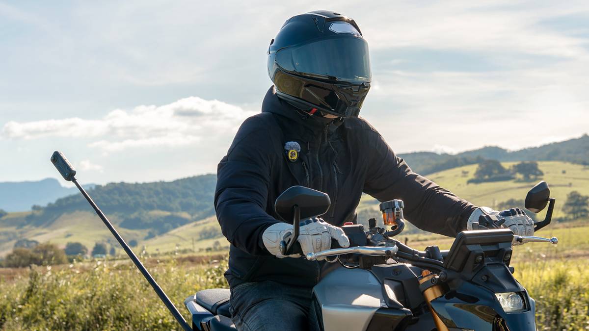 Motorcyclist in a black full-face helmet and black jacket with an Insta360 Mic transmitter clipped to his chest, riding a naked motorcycle with an Insta360 360 camera on a selfie stick through green hills