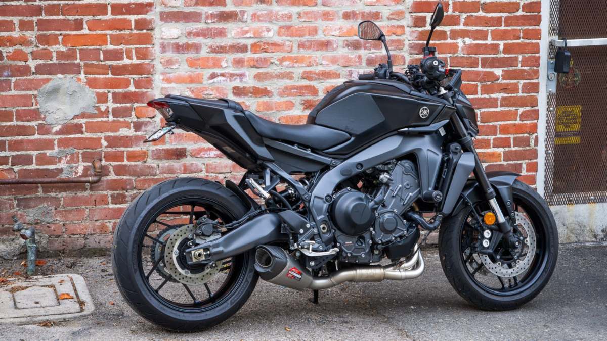 Black U.S.-spec Yamaha MT-09 fitted with the Yoshimura AT2 Speed Series stainless full exhaust and carbon end cap, parked against a red brick wall